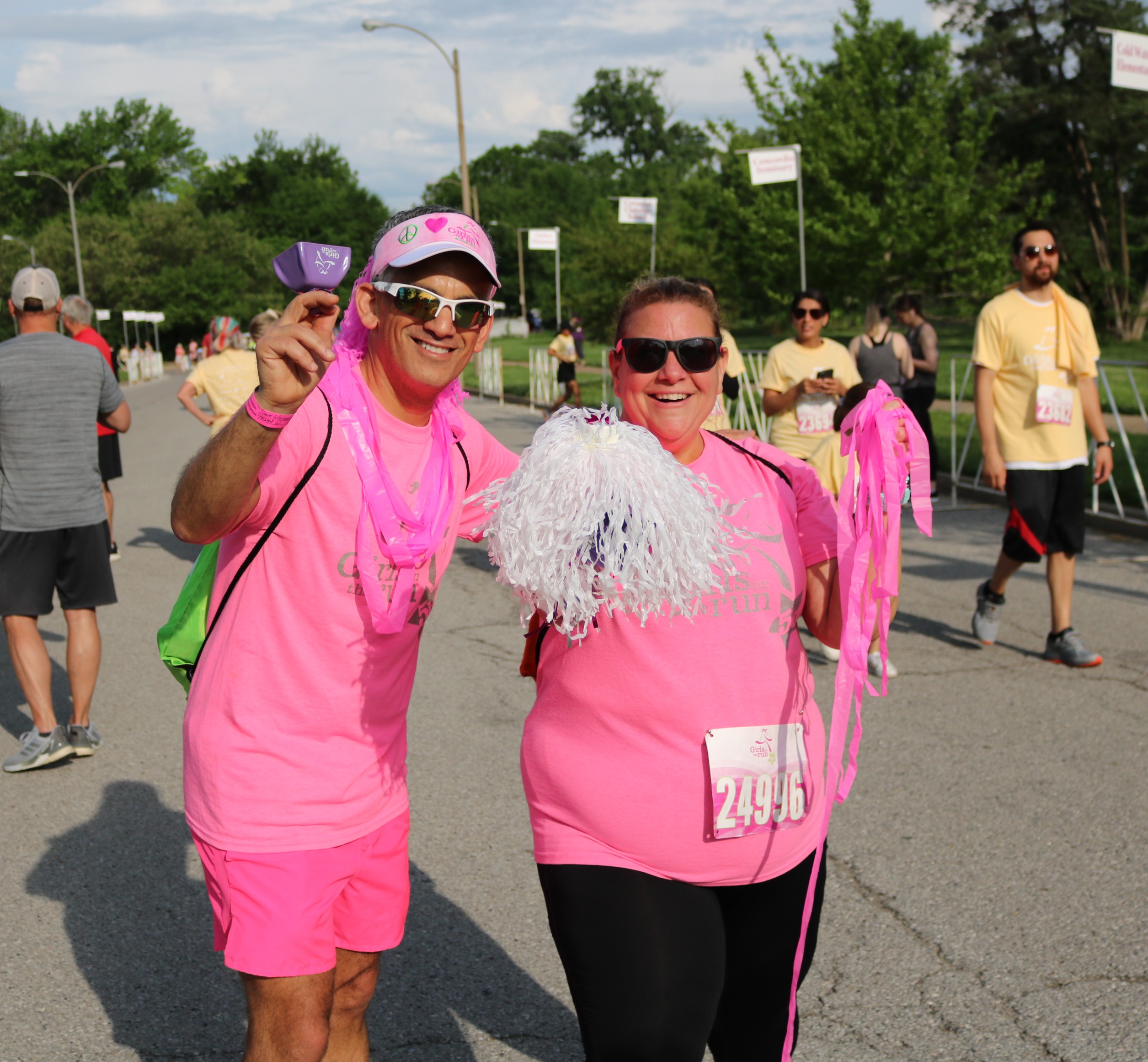 2 sparklers in bright pink shirts smile and wave a pom pom and cowbell at the 5K